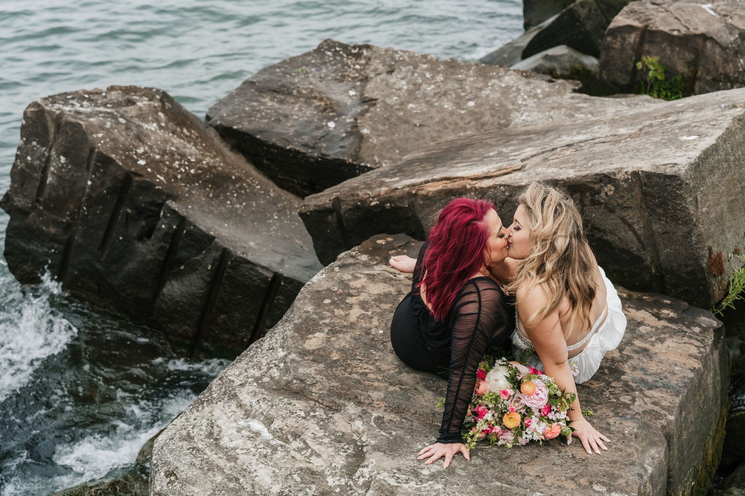 Same-sex bridal couple sits on rocks by the lake in Glacier National Park