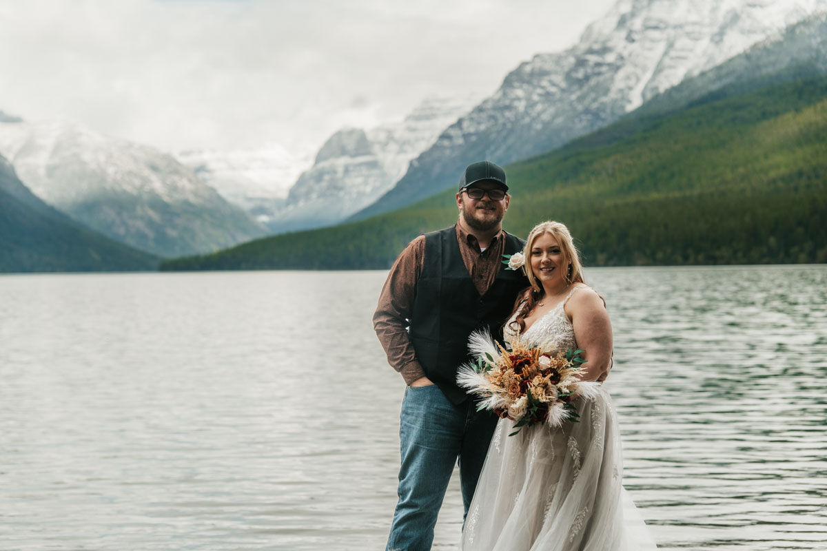 Montana elopement couple walking down the dirt road to Bowman Lake with tall pines and mountain peaks in the distance.
